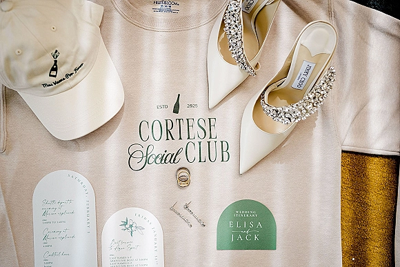 Wedding flat lay featuring a wedding invitation suite, itinerary card, rings, drop earrings, and bridal heels on textured fabric background