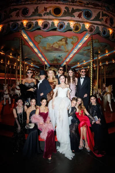 Wedding party portrait as the bride with friends gather by a glowing carousel with painted canopy, one wearing a chic feather wrap.