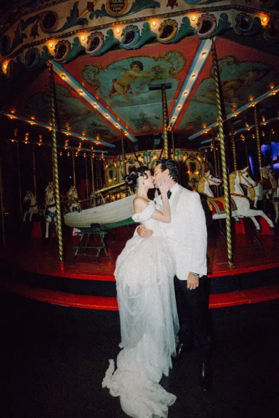 Wedding kiss portrait, a night wedding portrait: the couple kiss beneath glowing carnival lights on a carousel, her off-shoulder sleeves.