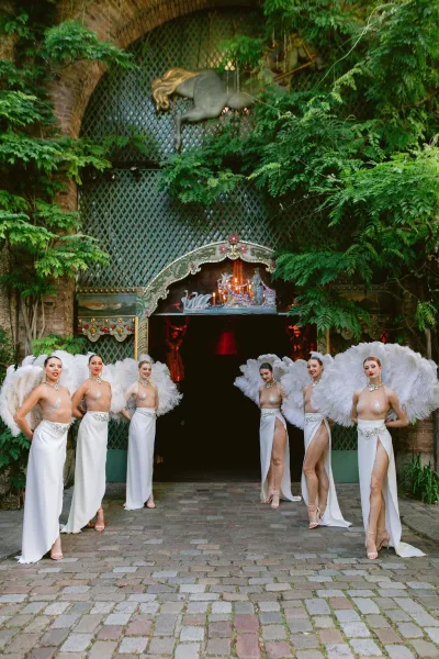 Wedding entertainment showgirl performers pose with sweeping feather fans under a brick archway in an ivy-lined cobblestone courtyard.