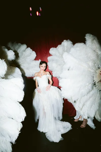 Bridal portrait in a feather bridal gown before a red curtain on a dark stage, softly lit by warm stage lights.