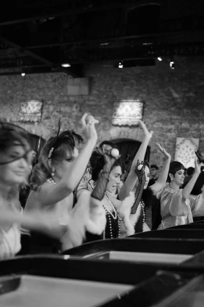 Reception party moment with lively wedding reception entertainment as flapper-clad guests cheer in feather headpieces by an exposed brick wall.