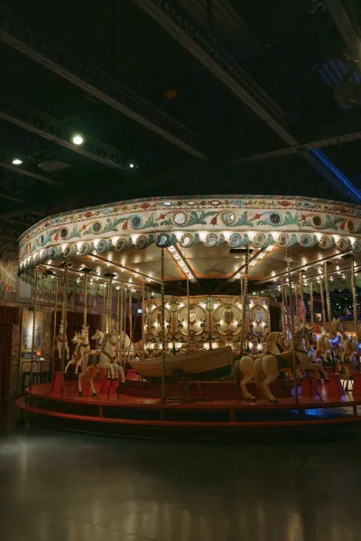 Vintage carousel with merry go round horses glows under marquee lights, set indoors against red curtains and wall murals beneath trusses.