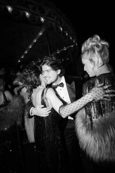 Reception moment as guests share wedding hugs, one in a sequin dress, beneath glowing carousel lights of a carnival ride at night.