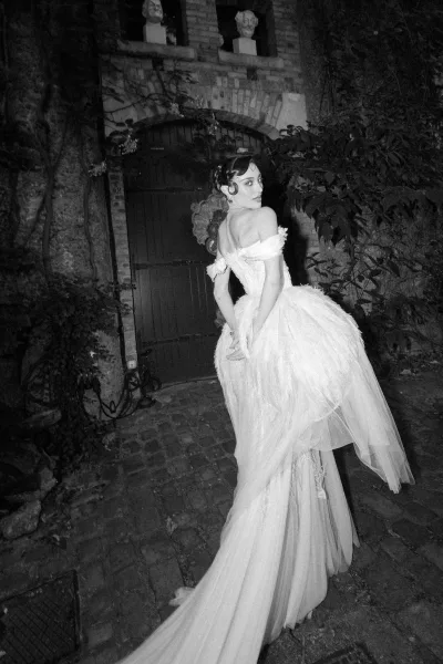 Bridal portrait of a bride in an off the shoulder wedding dress with a corset bodice and long train on a cobblestone path by an ivy-clad arch.
