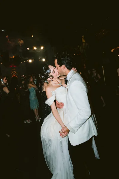Wedding kiss portrait as the couple share a dance floor kiss beneath string lights, her tulle train trailing while guests watch in the dark venue.