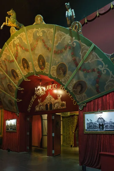 Carnival entrance decor featuring a vintage fairground arch and red curtains at a museum doorway with red walls and concrete floor.