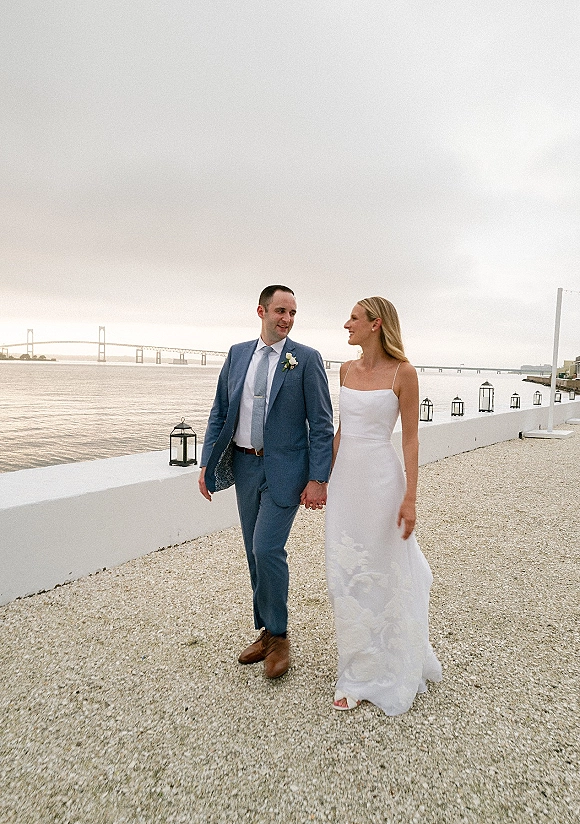Couple portrait of bride and groom walking hand in hand, her satin dress and his blue suit, by a waterfront bridge with lanterns