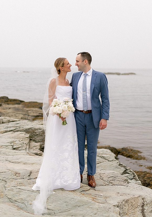 Couple portrait of bride and groom by ocean, smiling with white bouquet and veil on a rocky shoreline under overcast sky