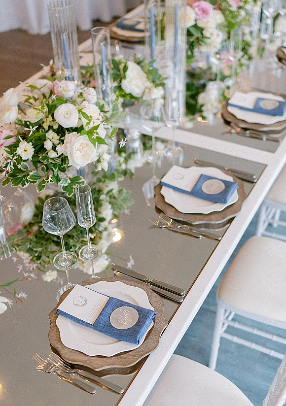 Reception tablescape with long head table decor, white and blush centerpiece, taper candles, blue napkins, scalloped plates on wood chargers and glassware