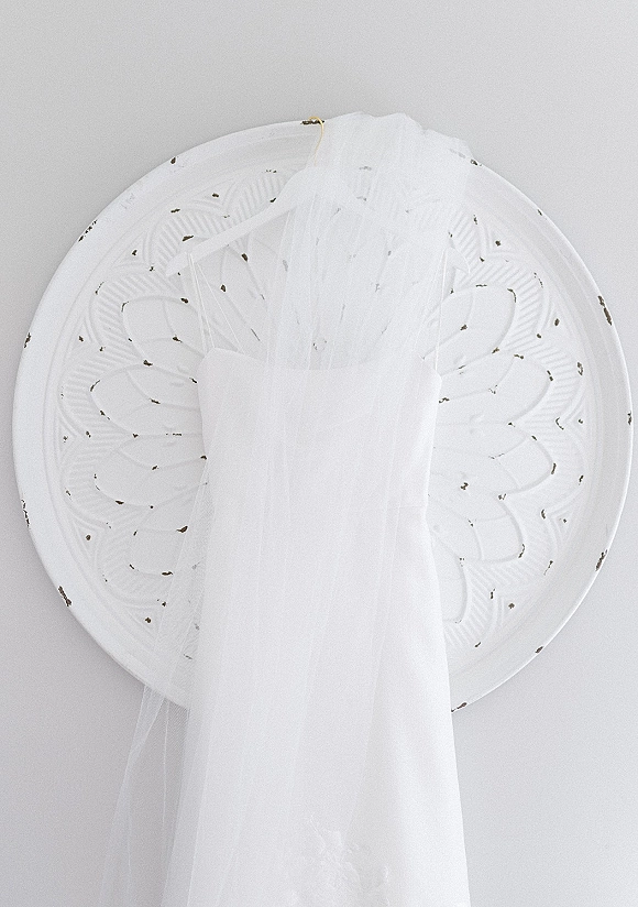 Wedding dress hanging on a hanger, a strapless wedding dress with lace applique and tulle veil accents against a light wall