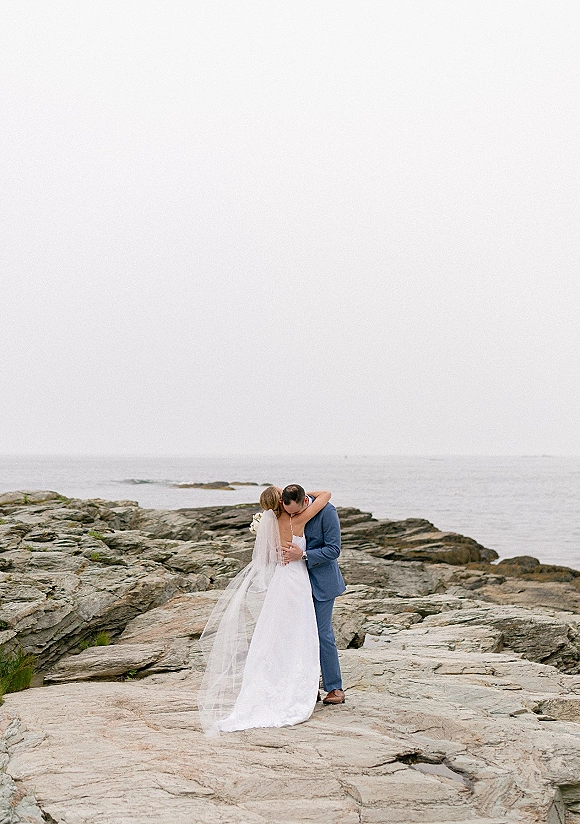 Wedding kiss portrait of bride and groom kissing, veil blowing, holding a white bouquet on a rocky ocean shoreline under overcast sky