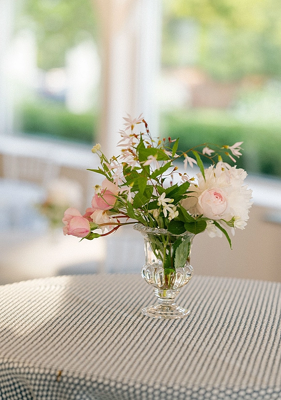 Wedding centerpiece with pink roses and white blooms in a clear glass vase, set on a striped tablecloth in soft window light