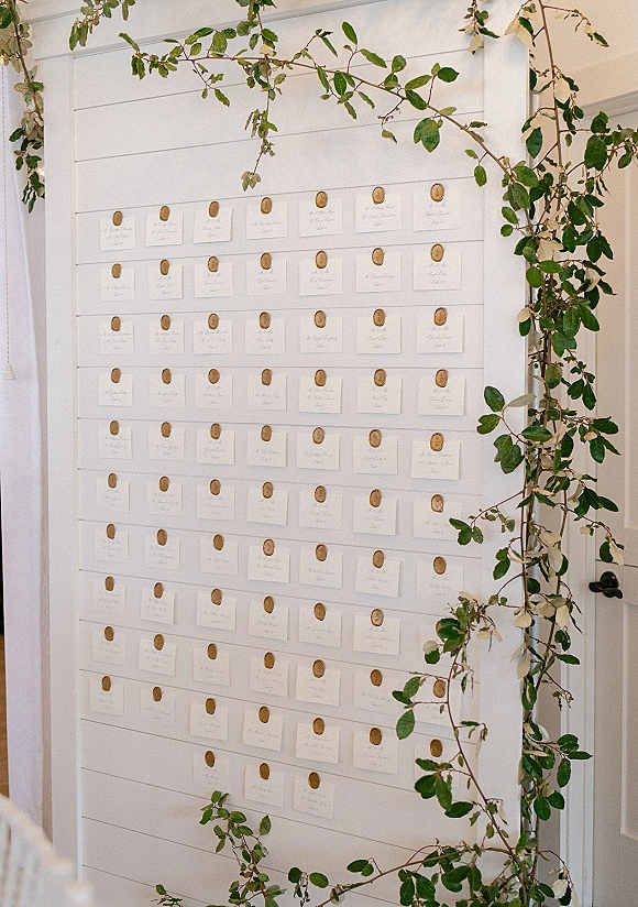 Wedding seating chart with escort card display using calligraphy cards and round wooden magnets, framed by greenery garland on a white shiplap wall