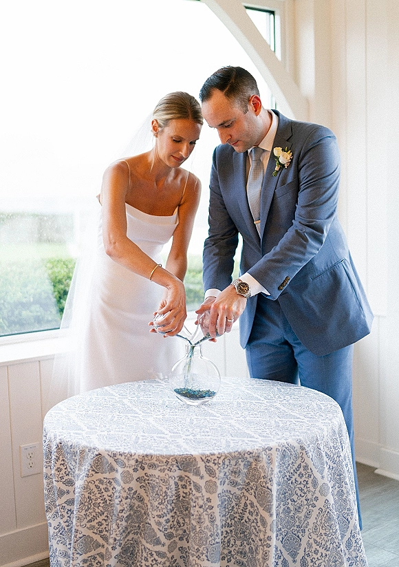 Unity ceremony as bride in a veil and groom in a suit pour colored sand into glass vases on a blue toile table by bright windows