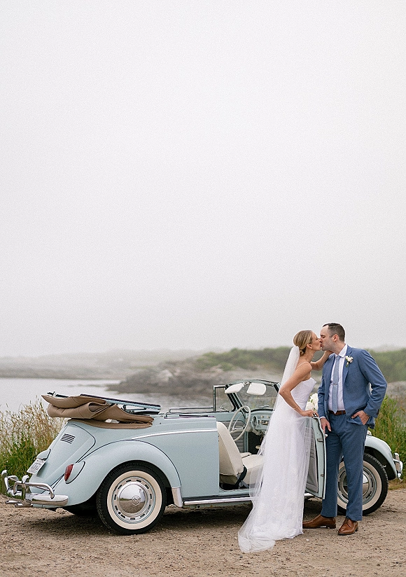 Wedding couple portrait of bride and groom kissing beside a vintage convertible, her veil lifting in ocean wind on coastal cliffs