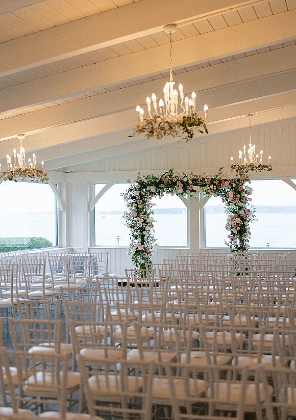 Indoor ceremony setup with wedding ceremony chairs facing a rose and greenery floral arch beneath chandeliers in a bright waterfront pavilion