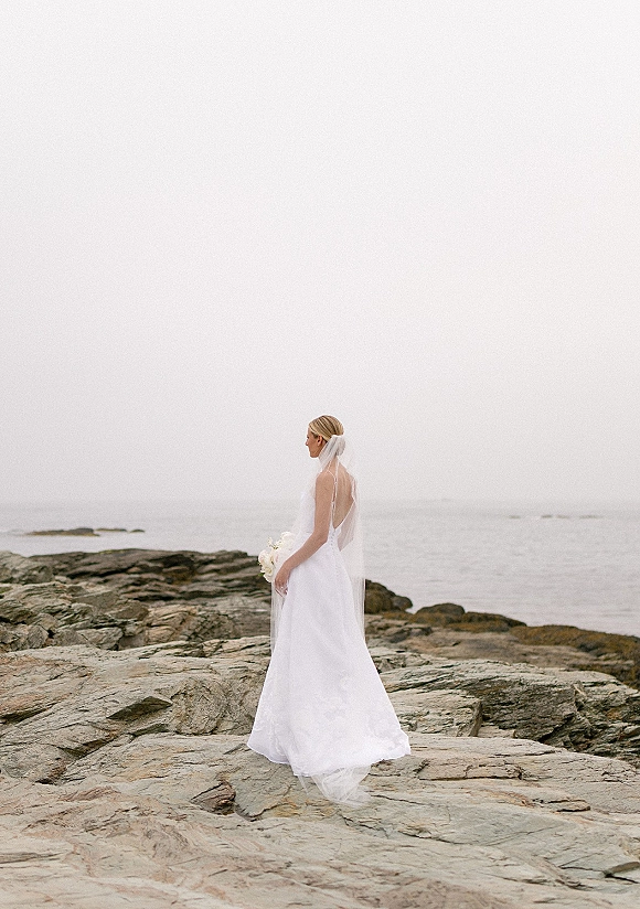 Bridal portrait of a bride in a wedding dress with veil holding a bouquet on a rocky shoreline, looking toward the ocean under overcast sky