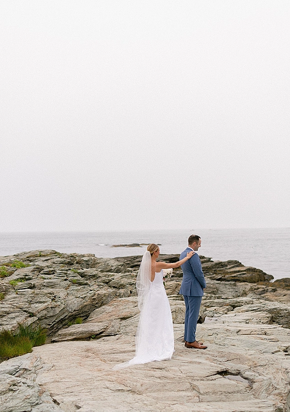 First look moment as the bride taps the groom’s shoulder, long veil and bouquet trailing on a rocky coastline under an overcast sky