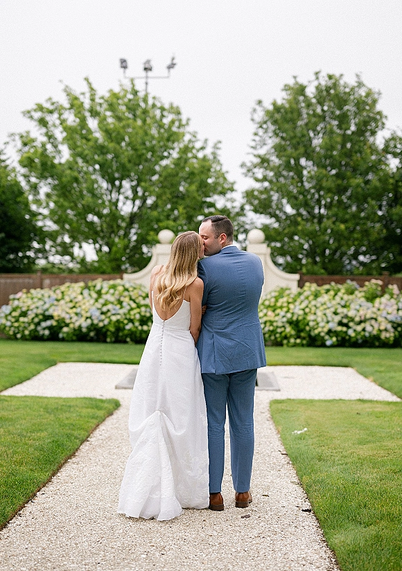 Wedding couple portrait of bride and groom from behind kissing on a gravel garden path, groom in blue suit under overcast sky