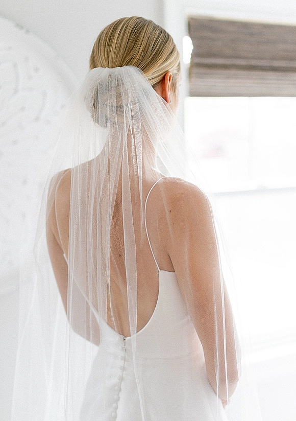 Bridal portrait of a bride from behind in a cathedral veil over a low bun, wearing a button-back spaghetti strap dress by window light indoors