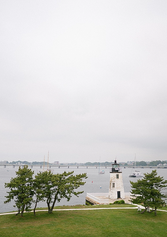 Lighthouse waterfront view with a white railing accent, sailboats and a buoy on calm harbor water under cloudy skies near a distant bridge