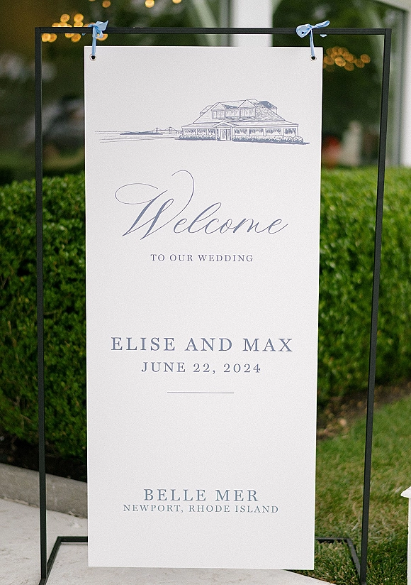 Wedding welcome sign hanging on a metal stand with blue ribbon bows and grommets, set against hedge greenery and bistro lights outdoors