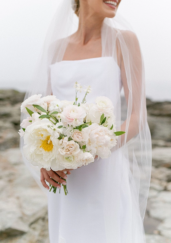 Bridal bouquet of white peonies and garden roses with greenery in a bride’s hands, veil and dress visible by a rocky ocean shore