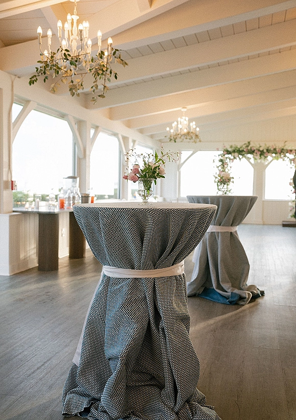 Wedding cocktail tables with grey linens tied in white bows, pink rose vase centerpieces, and chandeliers in a bright windowed room