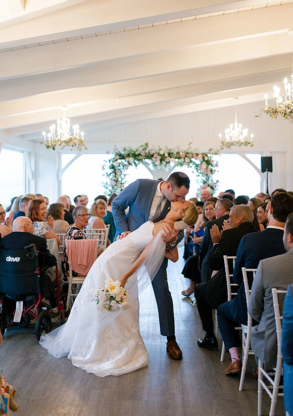 Wedding kiss portrait of groom dipping the bride as they kiss, veil and bouquet flowing down a bright indoor aisle under chandeliers