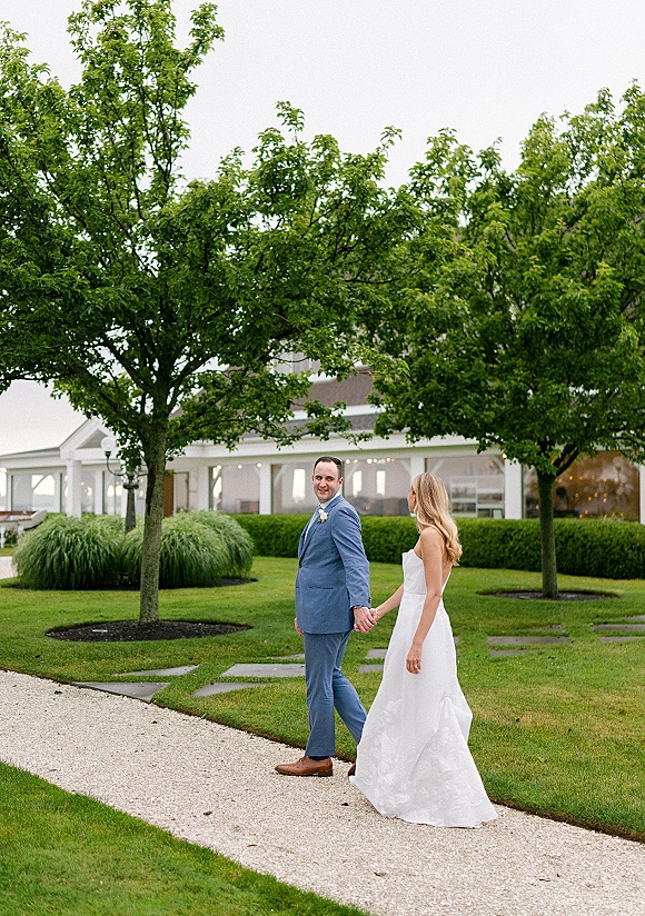 Couple portrait of bride and groom holding hands on a gravel path, looking back, with trees and a pavilion under an overcast sky