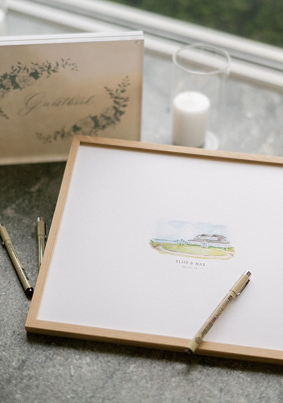 Wedding guest book in a wood frame with watercolor illustration and pens on a marble countertop by window light with greenery outside