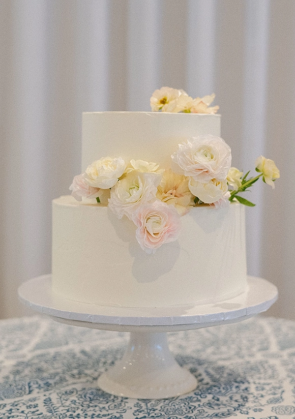 Wedding cake with white buttercream frosting, two-tier design, and fresh flowers on a stand atop a patterned tablecloth by white drapery