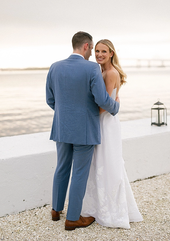 Couple portrait of a beach wedding couple embracing, bride looking over shoulder in strapless dress by ocean and shoreline wall with lantern
