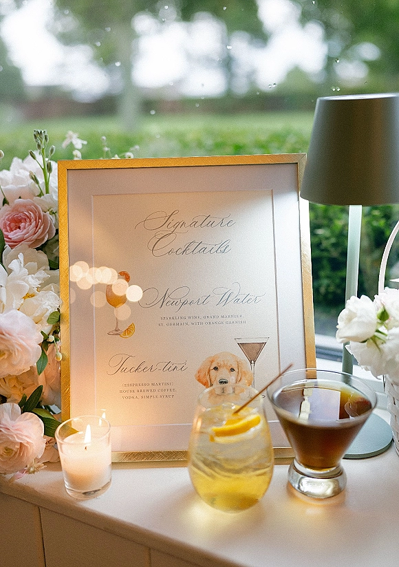 Signature cocktail sign in a gold frame with floral arrangement and cocktail glasses on a bar table by a rain-speckled window