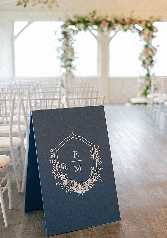Wedding welcome sign in navy with monogram and white ink floral crest beside white Chiavari chairs in an indoor ceremony space with windows