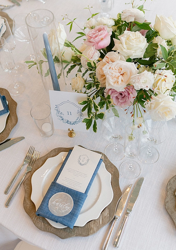 Reception tablescape with wedding place setting, rose and greenery centerpiece, blue taper candle, calligraphy place cards on white linens