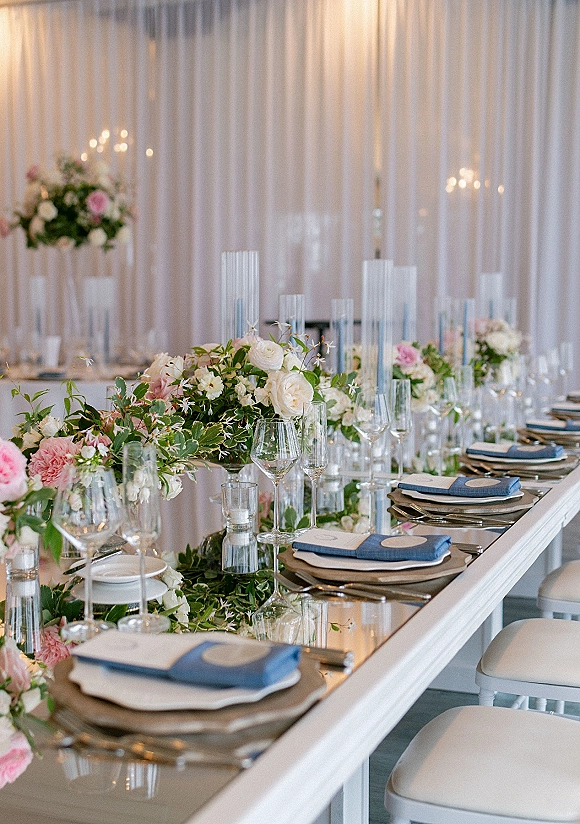 Reception tablescape with long banquet table decor featuring greenery garland, floral centerpieces, candles, and blue napkins under warm bistro lights