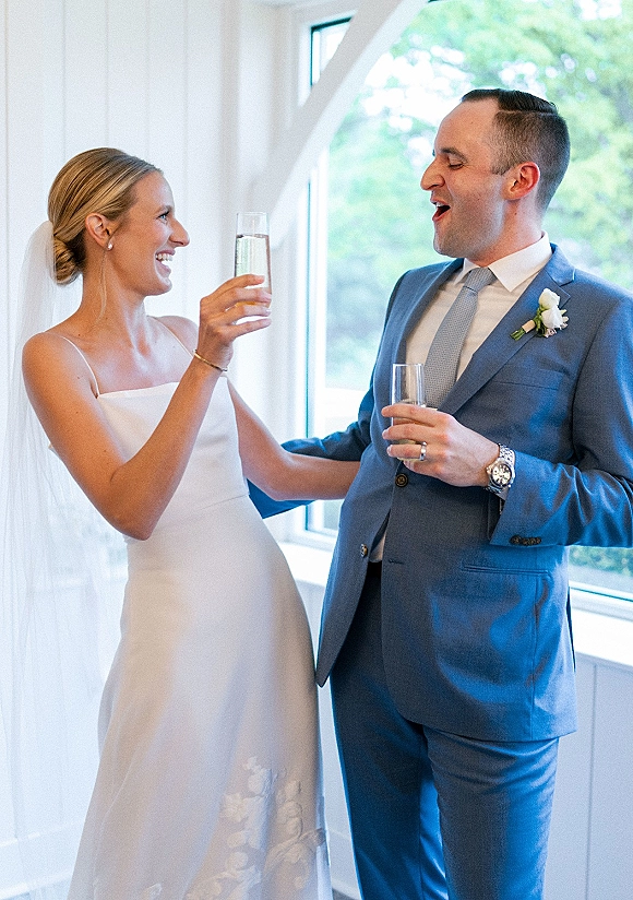 Wedding toast as bride and groom toasting with champagne flutes, smiling by an indoor window in veil and blue suit with boutonniere