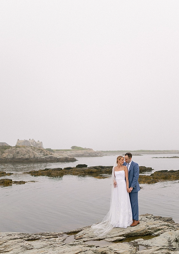 Couple portrait of bride in strapless wedding dress and veil with groom in blue suit on rocky shoreline by ocean and tide pools under overcast sky
