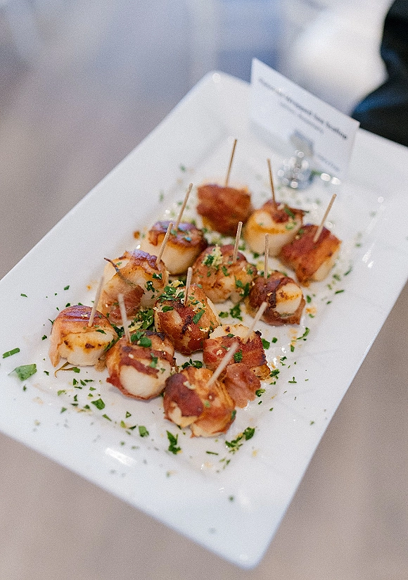Wedding cocktail appetizers featuring bacon wrapped scallops on a white rectangular platter with toothpicks and herb garnish on a neutral tabletop