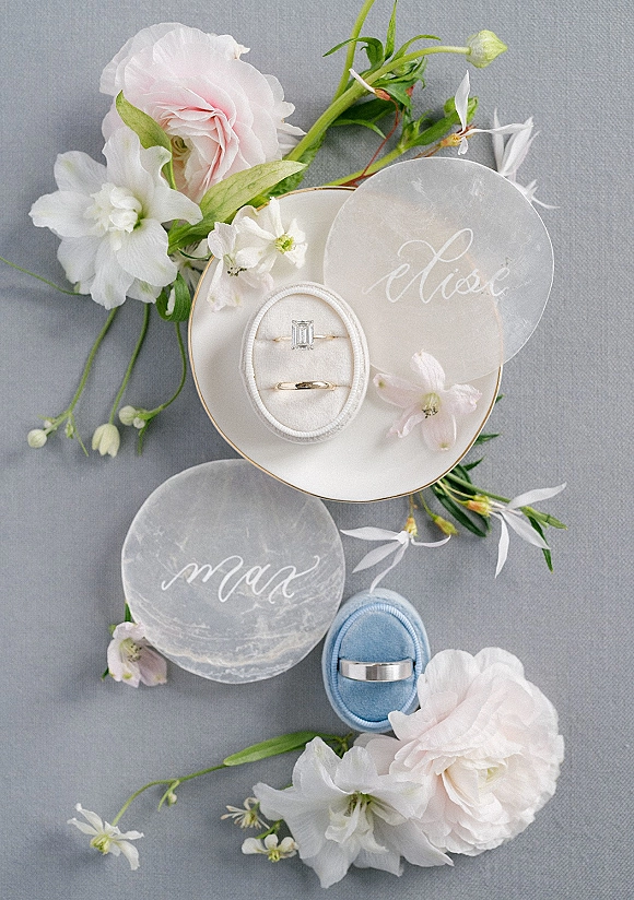 Wedding rings in a wedding ring flat lay on a white plate with gold rim, ring boxes, calligraphy name discs, and blush florals on gray fabric
