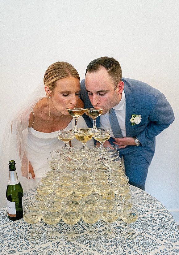 Champagne tower toast as bride in veil and groom in blue suit clink coupe glasses beside a champagne bottle on a round table