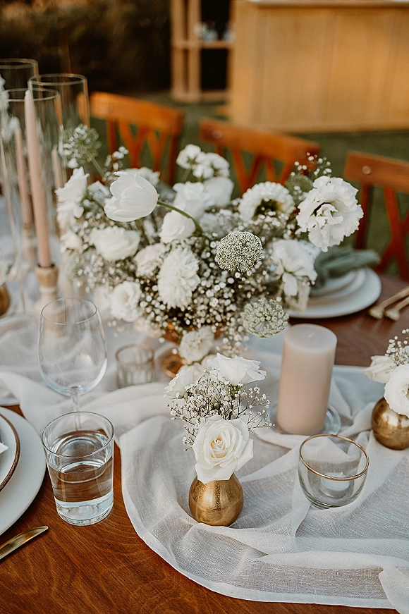 Reception tablescape with a wedding table centerpiece of white roses and baby’s breath, taper candles, gold bud vases, and linen runner on wood chairs backdrop