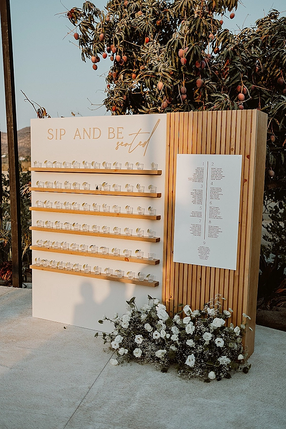 Seating chart display with wedding seating chart sign in calligraphy above escort shot glasses on wooden shelves, set on an outdoor patio backdrop
