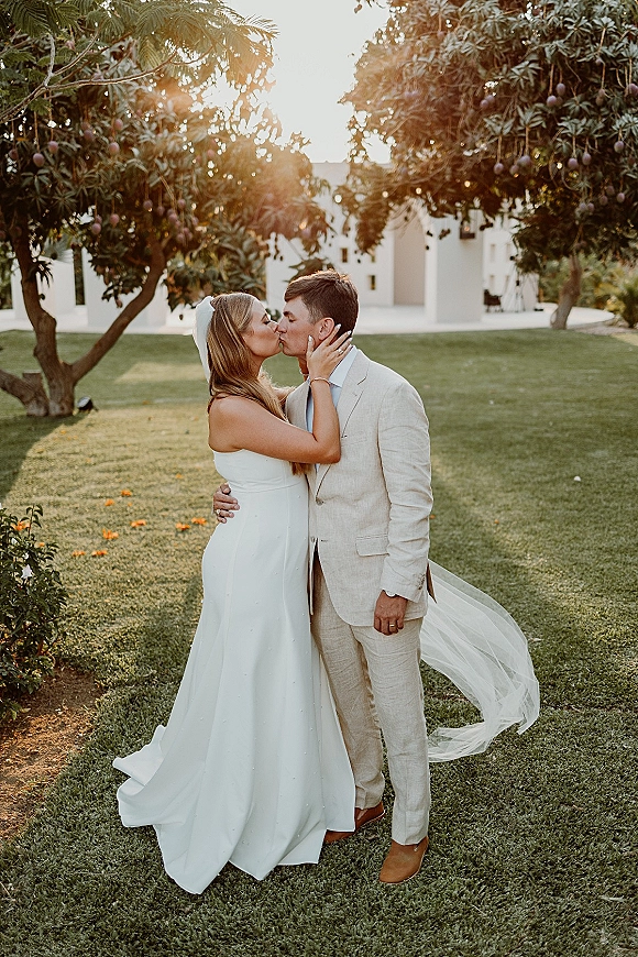 Wedding kiss portrait of bride and groom kissing as she holds his face, long veil blowing in sunlight on a lawn by a white building