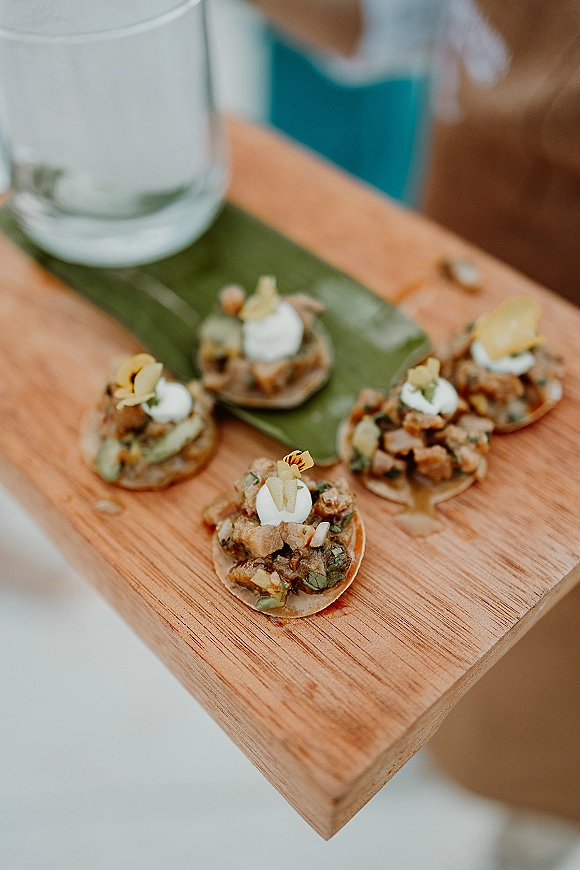 Wedding cocktail hour appetizers arranged as bite-size canapés on a wooden serving board with edible flower accent and blurred guest holding a drink glass