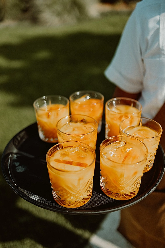 Wedding welcome drinks on a serving tray, orange cocktails with ice in glass tumblers carried by a server across a grassy lawn