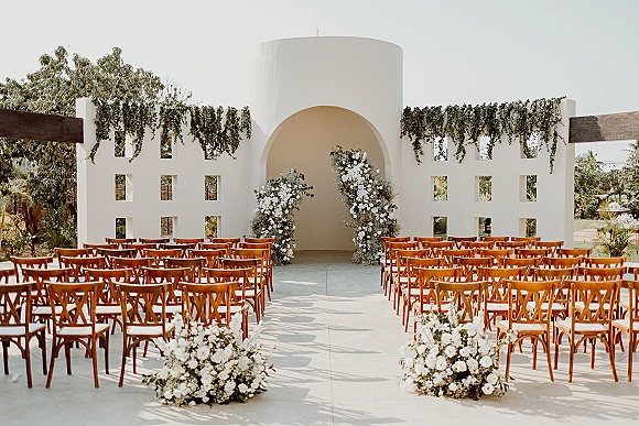 Ceremony setup with outdoor ceremony seating, wood crossback chairs lining an aisle to a white floral arch in a stucco courtyard alcove