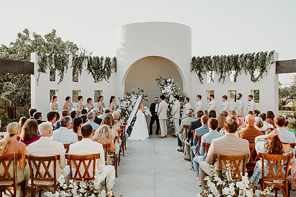 Wedding ceremony wide shot of bride and groom at altar with officiant, bridal party, and white floral arch in courtyard under stucco arch wall